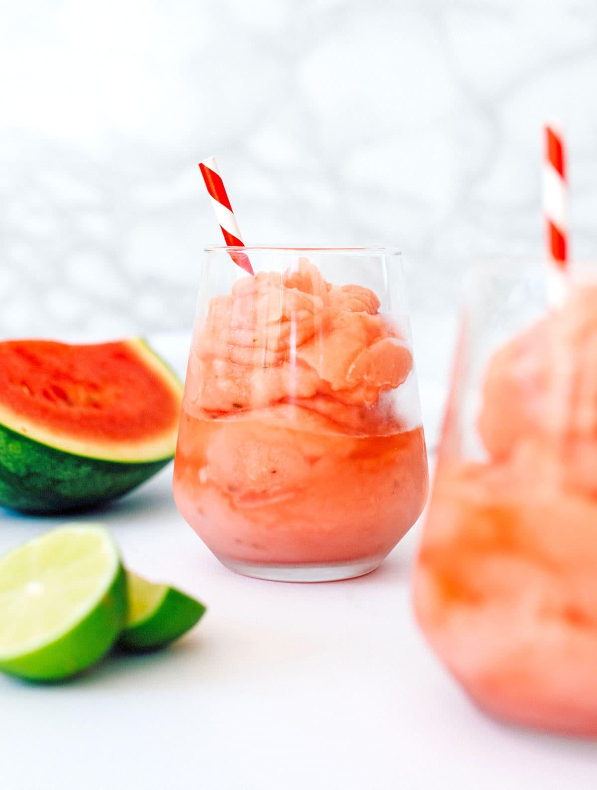 Gefrorener Wassermelonen-Daiquiri 2 Wassermelonen-Daiquiri in einem Glas mit rot-weißem Strohhalm und Wassermelonen- und Limettenscheiben daneben.