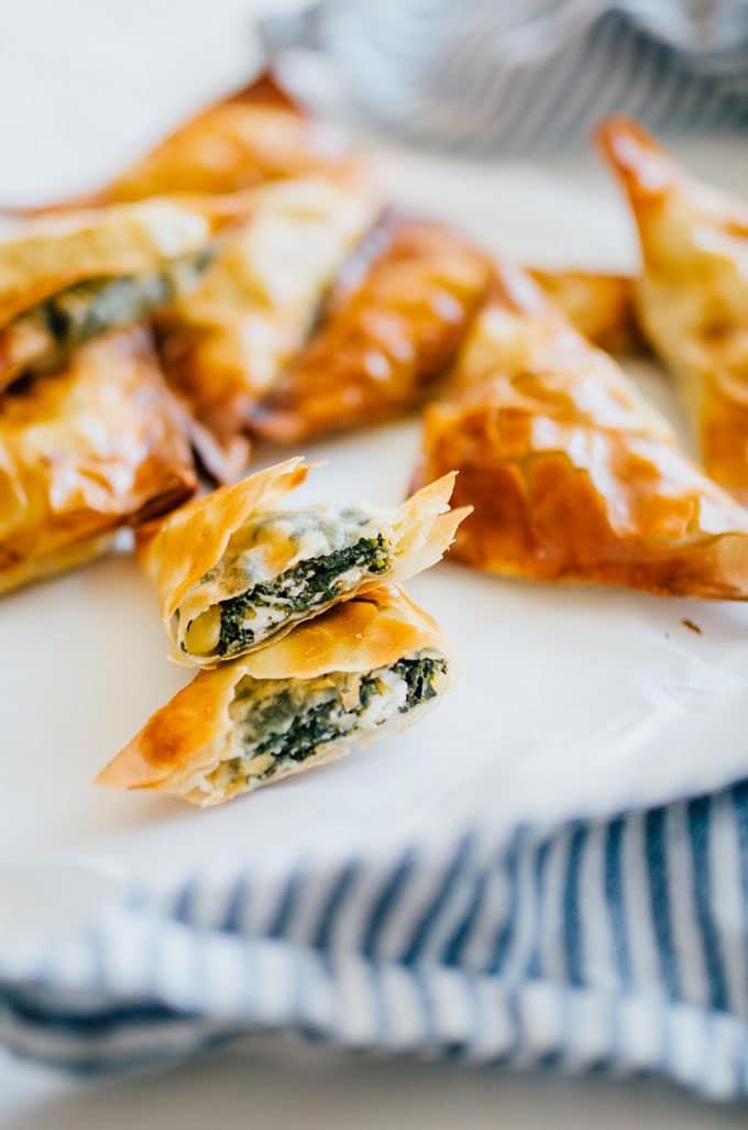 Spanakopita-Dreiecke (Kleine griechische Spinattörtchen!) 2 Wie man Spanakopita zubereitet, ein aufgeschnittenes Spanakopita Dreieck auf einem weißen Teller mit blauem Handtuch – Diese Spanakopita Dreiecke sind eine einfache Vorspeise, vollgepackt mit Feta und Spinat, umhüllt von butterigem, blättrigem Filoteig.