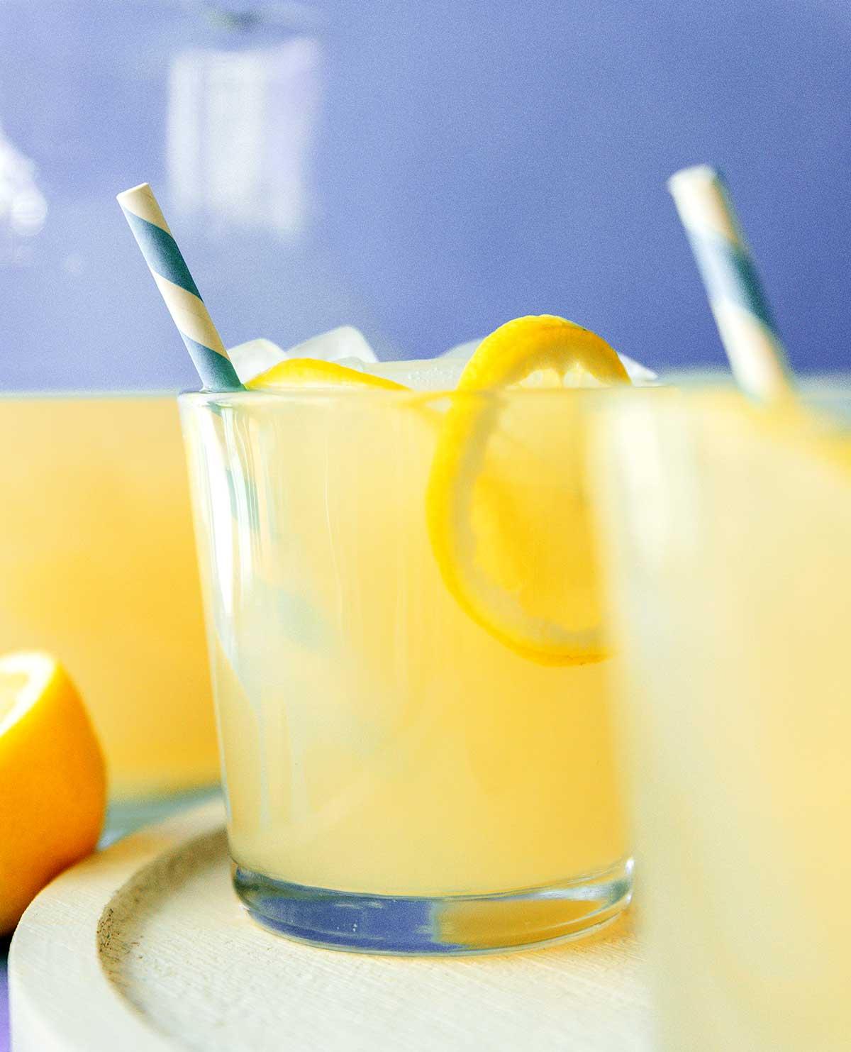 Einfache Lavendel-Zitronenlimonade (Saft oder Slush!) 2 Zwei Gläser Lavendel-Zitronenlimonade, verziert mit Zitronenscheiben und essbaren Blüten, stehen auf einem Holztisch, im Hintergrund sanft verschwommenes Lavendelfeld.
