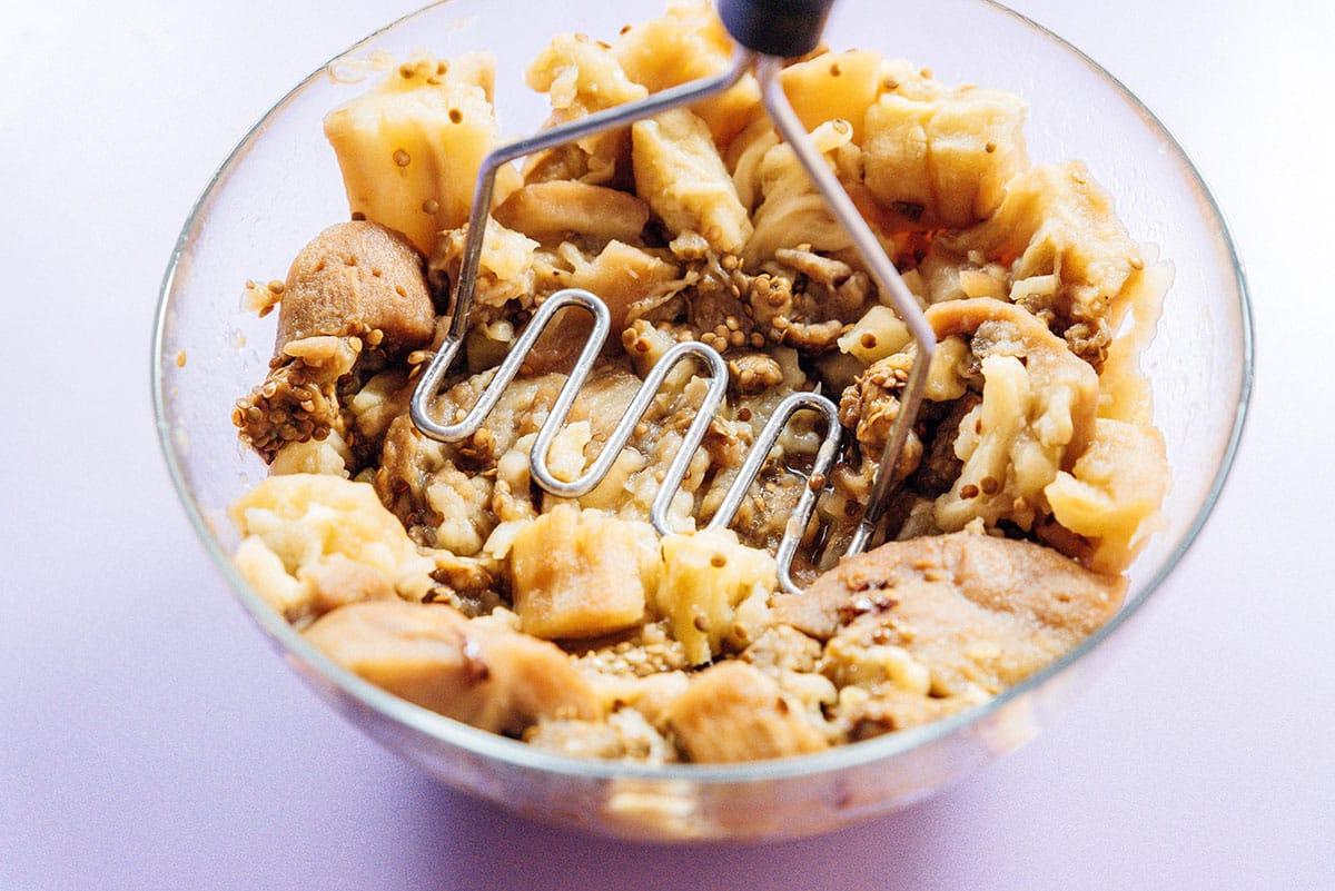 Griechischer Auberginen-Dip (Melitzanosalata) 11 Geröstete Aubergine wird mit einem Kartoffelstampfer in einer Schüssel zerstampft.