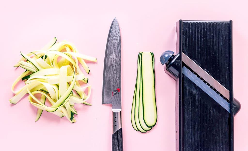 Zucchininudeln zubereiten 11 Ein Mandolinenschneider, 4 der Länge nach geschnittene Zucchinischeiben, ein Messer und ein Stapel Fettuccine-Zoodles auf rosafarbenem Hintergrund.