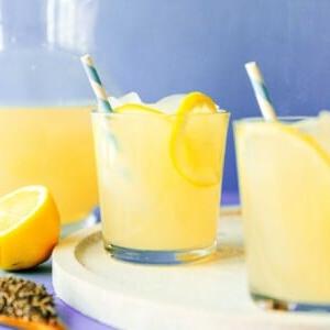 Einfache Lavendel-Zitronenlimonade (Saft oder Slush!) 11 Zwei Gläser erfrischender Limonaden-Slushie mit Strohhalmen und Zitronenscheiben stehen auf einem runden Brett, daneben eine halbierte Zitrone und ein Krug, vor einem leuchtend violetten Hintergrund.