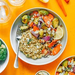 Geröstete Gemüse-Getreide-Bowls 13 Eine Getreide-Bowl mit Quinoa, geröstetem Rosenkohl, Karotten, roten Zwiebeln und Mandeln, garniert mit Zitronenspalten und Feta-Käse. Eine Gabel liegt daneben auf einem Hintergrund, der mit verstreutem Gemüse und Zitrone gesprenkelt ist.