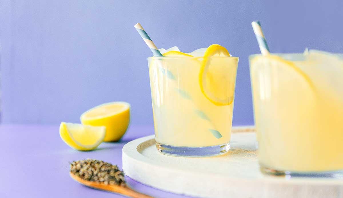 Zwei Gläser Limonaden-Slushie mit Strohhalmen und Zitronenscheiben stehen auf einem runden Tablett. Daneben liegen eine halbierte Zitrone und ein Löffel mit Kernen, vor einem violetten Hintergrund.