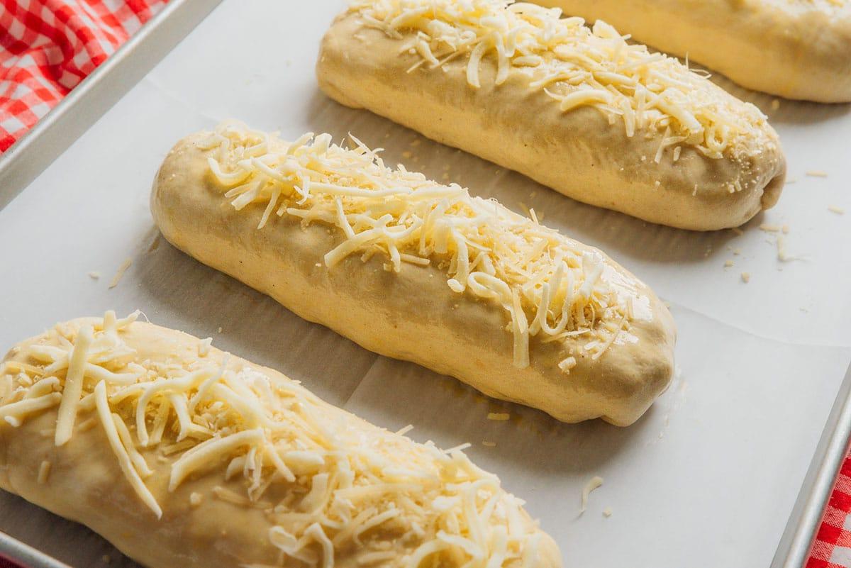 Vier ungebackene Brotlaibe, belegt mit geriebenem Käse, in einer Reihe auf einem mit Backpapier ausgelegten Backblech, auf einem karierten rot-weißen Tuch.