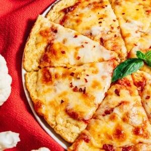 Blumenkohl-Pizzaboden zubereiten 14 Eine Käse-Pizza mit Blumenkohl-Pizzaboden wird auf einem Teller geschnitten, umgeben von frischem Blumenkohl und einer Schüssel Marinara-Sauce auf einem roten Tuch.