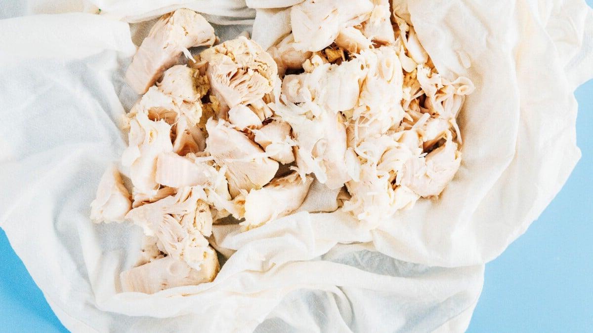 Zerkleinerte gekochte Hähnchenbruststücke auf einem weißen Tuch vor hellblauem Hintergrund, die der Textur von BBQ Jackfrucht Pulled Pork ähneln.