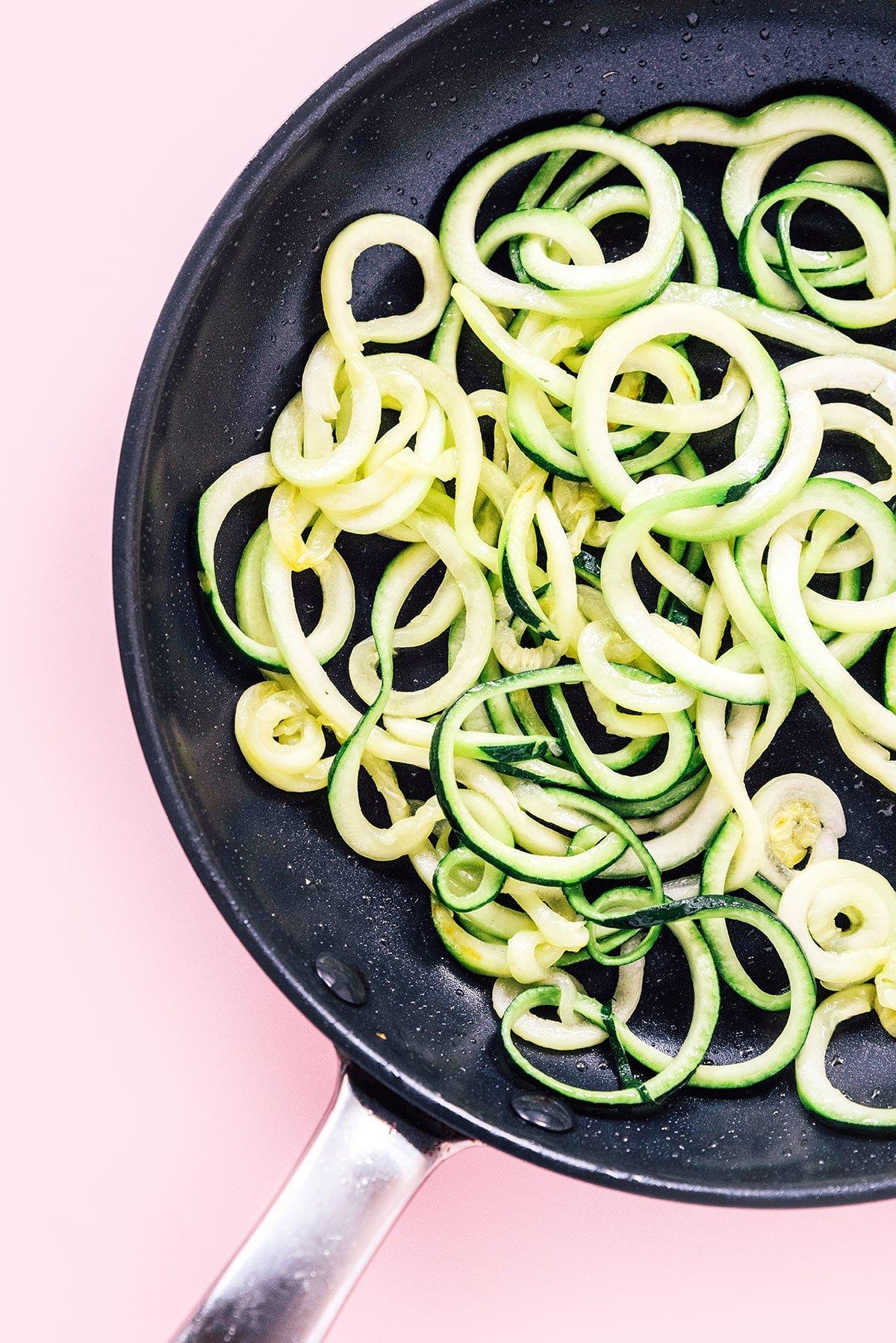 Zucchininudeln zubereiten 15 Eine schwarze Pfanne mit Spaghetti-Zoodles auf rosafarbenem Hintergrund.