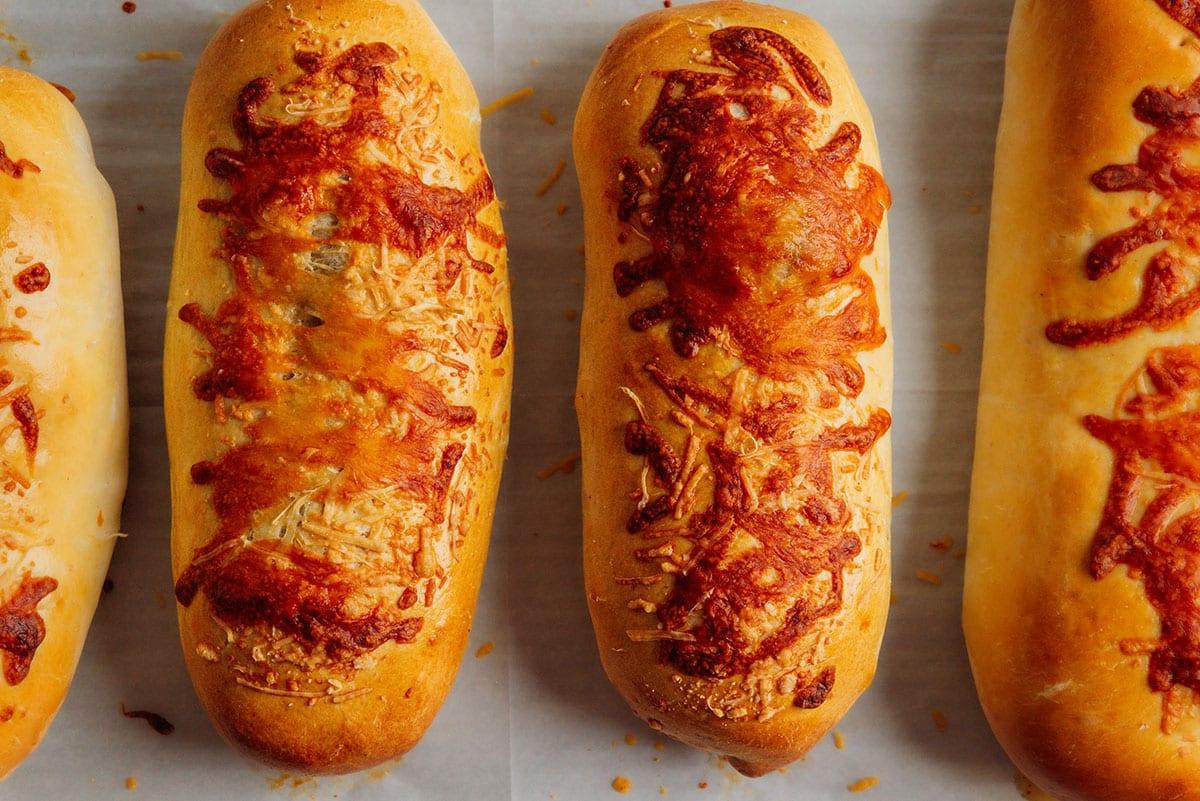 Vier goldbraune Brotlaibe mit geschmolzenem Käse, in einer Reihe auf Backpapier angeordnet.