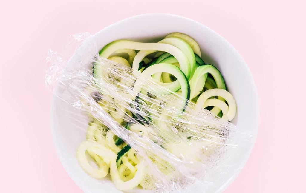 Zucchininudeln zubereiten 16 Eine weiße Schüssel auf rosafarbenem Hintergrund, gefüllt mit Spaghetti-Zoodles und teilweise mit Frischhaltefolie bedeckt. Als ob die Schüssel mit Frischhaltefolie bedeckt wird.