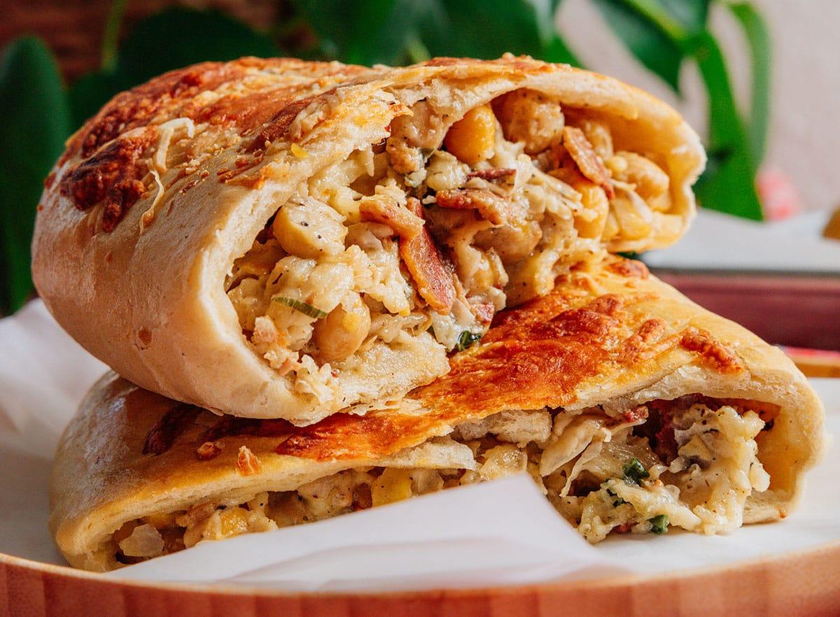 Nahaufnahme eines geschnittenen Burritos, gefüllt mit Kichererbsen, zerkleinertem Hähnchen und Kräutern, erinnert an eine hausgemachte Variante des beliebten Costco Chicken Bake. Er ist kunstvoll auf einem Holzteller arrangiert, mit einer verschwommenen grünen Pflanze im Hintergrund.