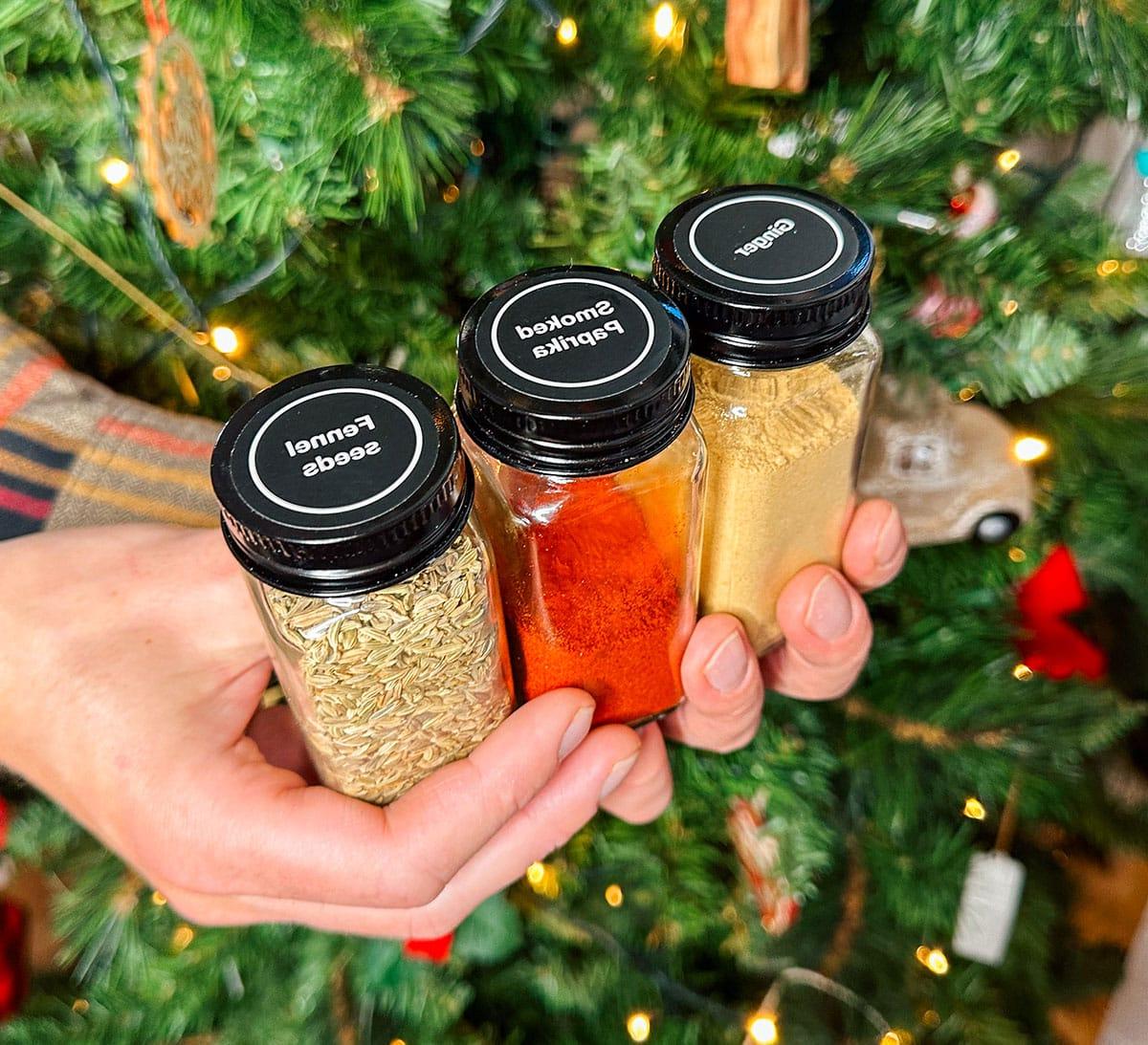 Beschriftete Gewürzgläser mit passenden Messlöffeln als Geschenk vor einem Weihnachtsbaum.