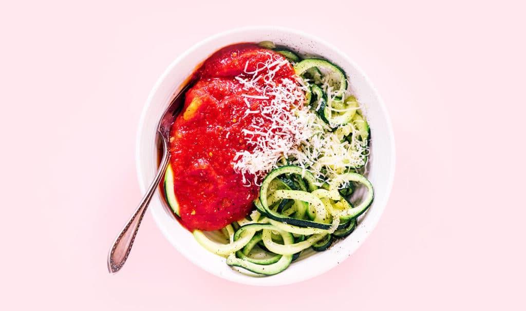 Zucchininudeln zubereiten 17 Spaghetti-Zoodles, rote Soße, Parmesan und eine Gabel in einer weißen Schüssel auf rosafarbenem Hintergrund.