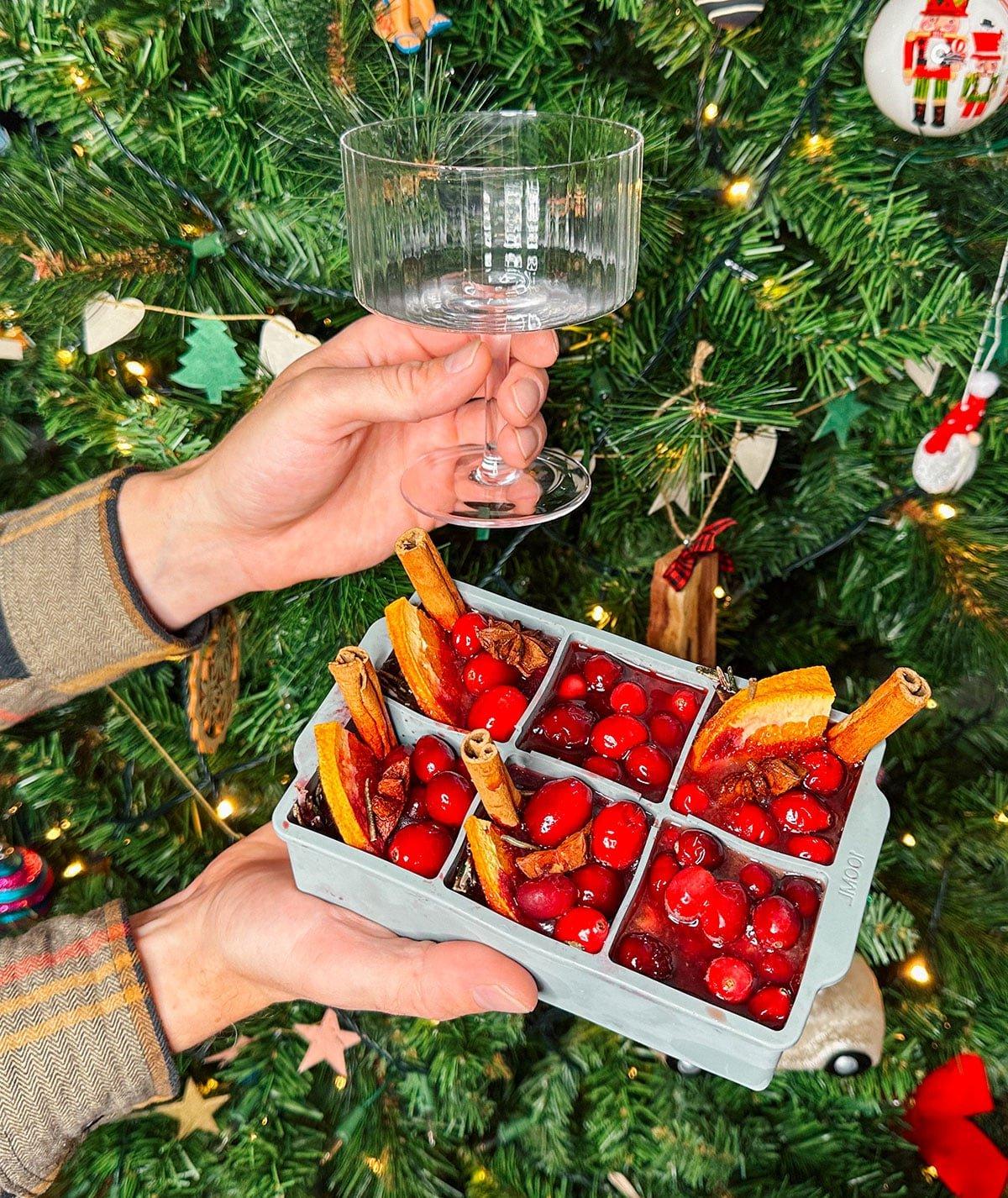 Riesige Eiswürfel in Cocktailgläsern (Cranberry Aperol Spritz) als Geschenk vor einem Weihnachtsbaum.
