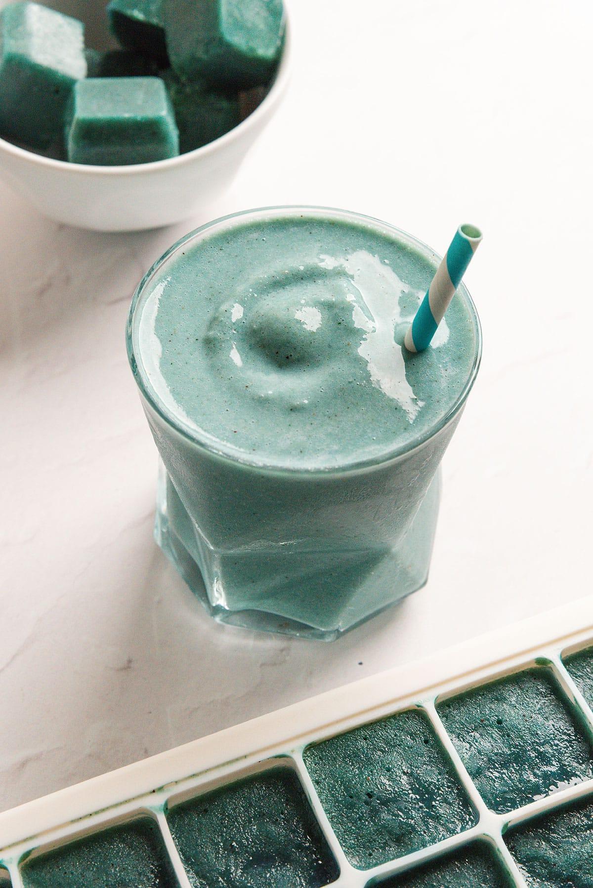 Smoothie-Würfel (Evive-Kopie) 19 Ein Glas dicker blassgrüner Smoothie mit einem gestreiften Strohhalm steht neben einer Schüssel und einem Tablett mit grünen Smoothie-Würfeln auf einer weißen Oberfläche.