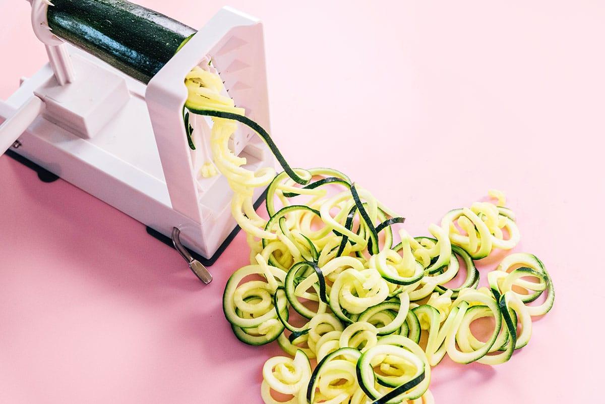 Entdecken Sie, wie Sie Zucchini-Nudeln mühelos mit einem Spiralschneider zubereiten und perfekte, gelockte Stränge auf einer leuchtend rosa Oberfläche kreieren.