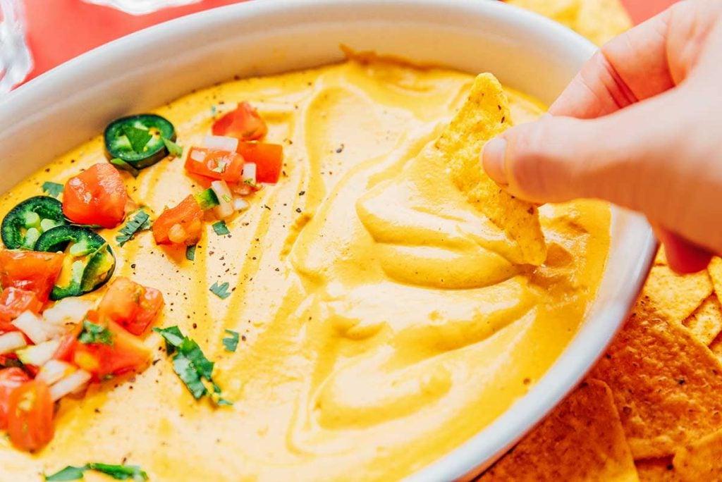 **10-Minuten-Rezept für veganen Queso** 3 Eine Hand taucht einen Tortillachip in eine Schüssel mit cremigem veganem Queso, garniert mit Jalapeño-Scheiben, Tomaten und frischem Koriander.