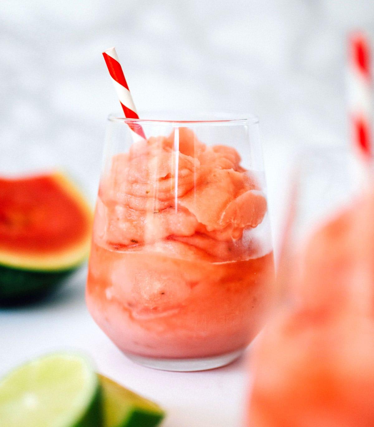 Gefrorener Wassermelonen-Daiquiri 3 Wassermelonen-Daiquiri in einem Glas mit rot-weißem Strohhalm und Wassermelonen- und Limettenscheiben daneben.