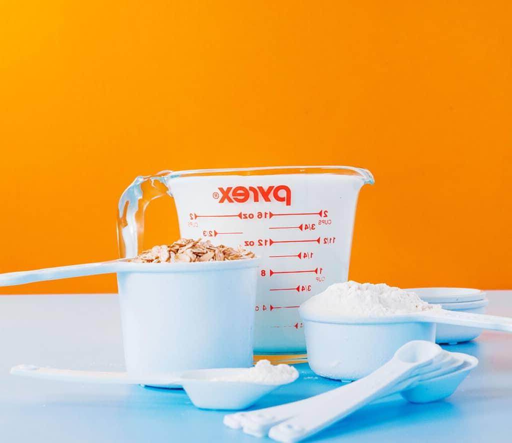 Wie viele Esslöffel sind in einer Tasse? (+ Umrechnungshilfe!) 3 Weiße Messbecher mit trockenen Lebensmitteln neben einem vollen Glasmessbecher mit Milch.