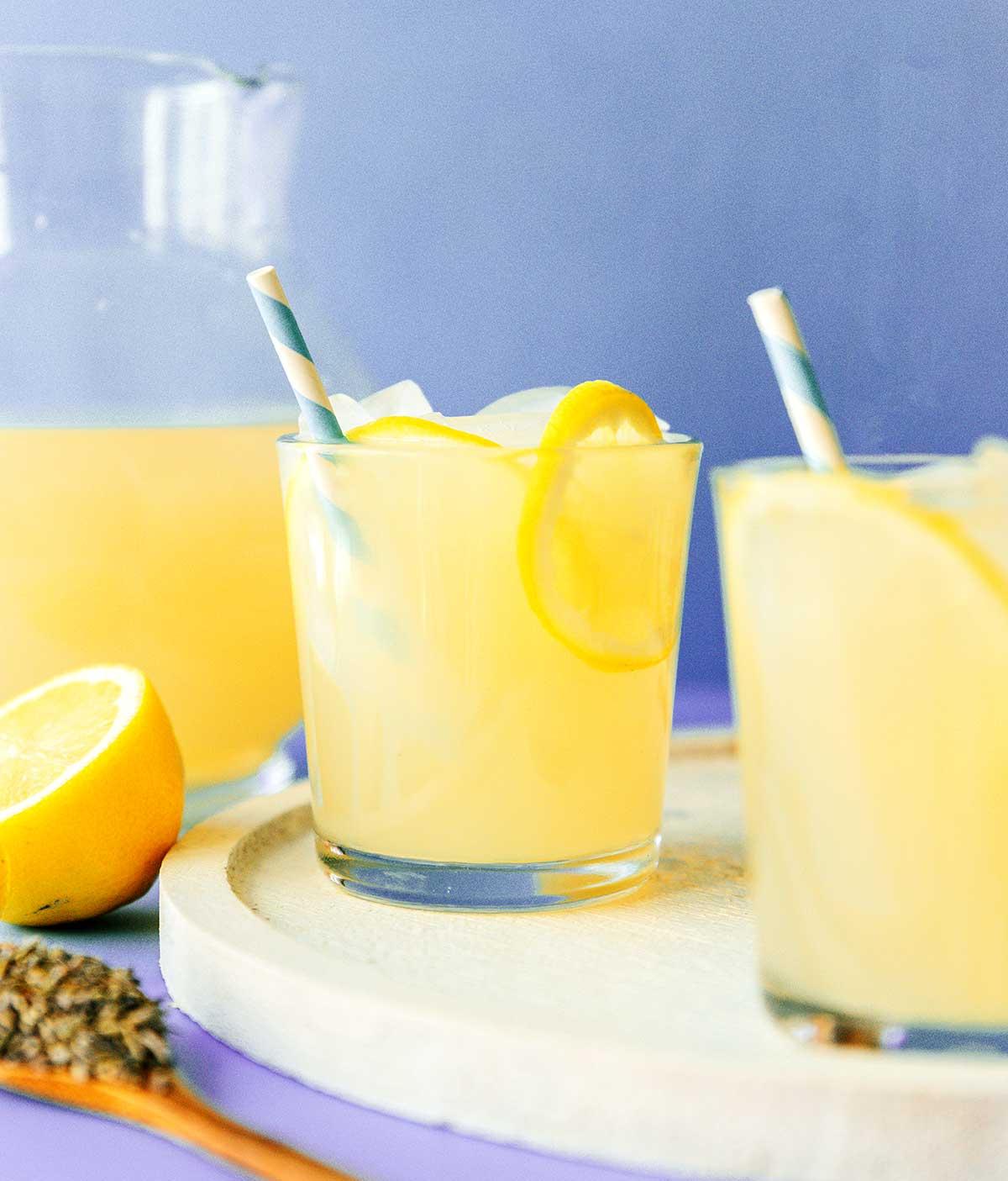 Einfache Lavendel-Zitronenlimonade (Saft oder Slush!) 3 Ein Glas Lavendel-Zitronenlimonade, verziert mit frischen Zitronenscheiben und einem Lavendelzweig, steht auf einem rustikalen Holztisch, im Hintergrund verschwommene Sommerlandschaft.