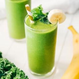 Ein hohes Glas Kale-Smoothie, garniert mit einer Bananenscheibe und einem Grünkohlblatt, komplett mit einem gestreiften Strohhalm. Ein weiteres Glas, frische Banane und leuchtende Grünkohlblätter erscheinen im Hintergrund.