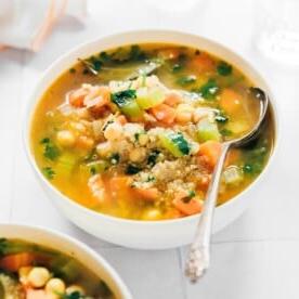 A bowl of quinoa vegetable soup with chickpeas, garnished with fresh herbs. A spoon rests on the bowls edge, while another bowl is partially visible in the background.