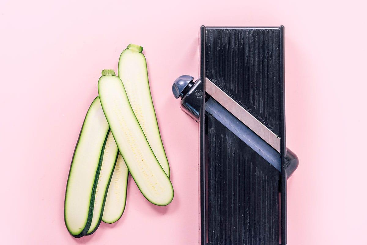Ein Küchenmandolinenschneider sitzt neben drei dünn geschnittenen Zucchinistücken auf einem rosa Hintergrund, perfekt für diejenigen, die herausfinden möchten, wie man Zucchini-Nudeln zubereitet.