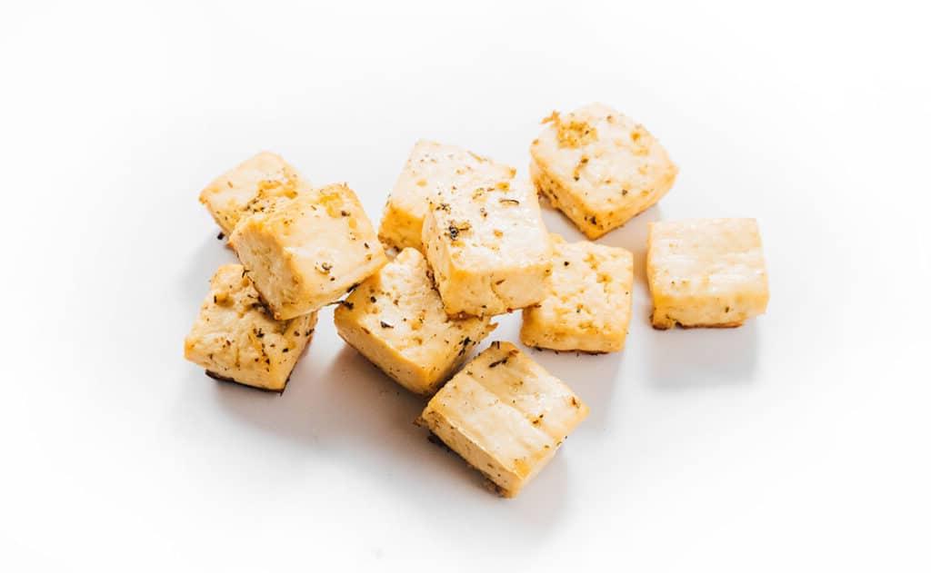 Wie man Tofu mariniert (8 Geschmacksrichtungen!) 24 Gebackene Knoblauch- und Kräuter-Tofu-Würfel auf einer weißen Oberfläche.