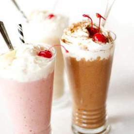 Three milkshakes, reminiscent of how to make nice cream, topped with whipped cream and cherries, stand tall in glasses with straws. Indulge in the classic flavors of chocolate, vanilla, and strawberry.