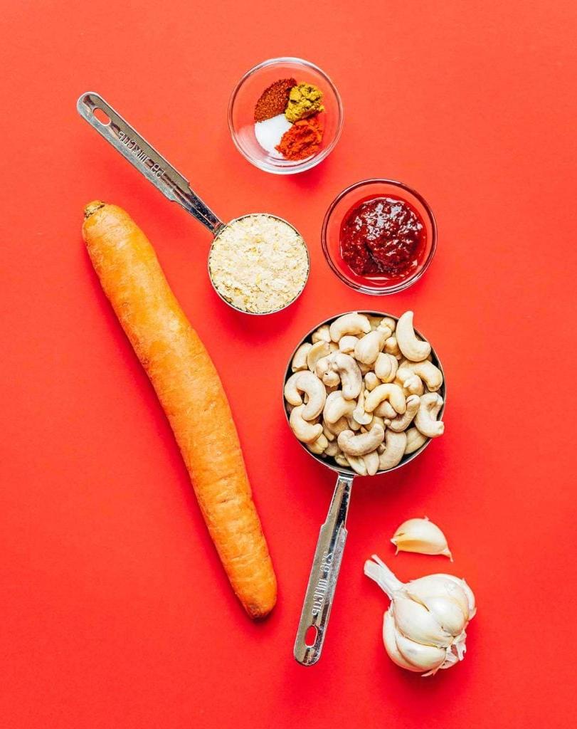 **10-Minuten-Rezept für veganen Queso** 4 Verschiedene abgemessene vegane Queso-Zutaten, wie Cashewkerne, Karotten, Gewürze und scharfe Sauce, auf einem roten Hintergrund arrangiert.
