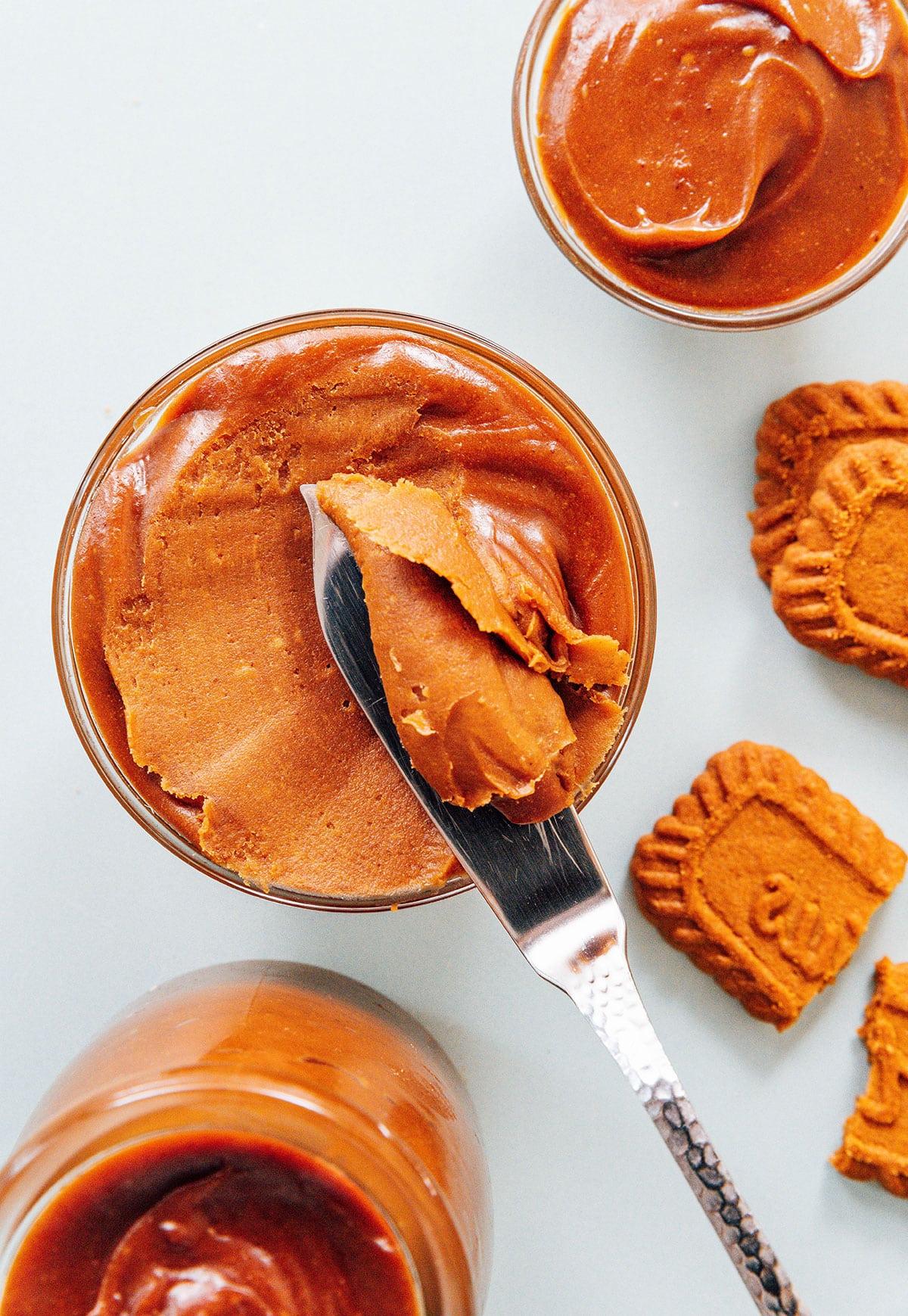 Wie man Spekulatius-Kekscreme herstellt 4 Zutaten für hausgemachte Keksbutter auf einer Arbeitsfläche, mit einem Glas Keksbutter im Hintergrund.