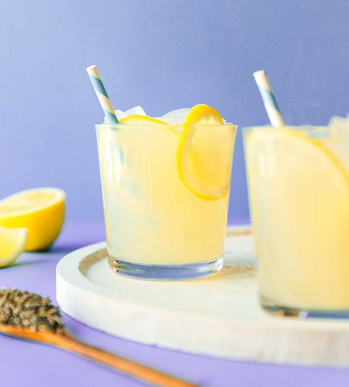 Einfache Lavendel-Zitronenlimonade (Saft oder Slush!) 4 Ein klares Glas mit Lavendel-Zitronenlimonade, verziert mit frischen Zitronenscheiben und essbaren Blüten, die auf der Oberfläche schwimmen, steht auf einem hölzernen Untersetzer.