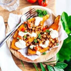 **10 Arten, Hummus zu essen (+ Rezepte!)** 4 Mediterran gefüllte Süßkartoffeln, serviert als gebackene Hälften, garniert mit gerösteten Kichererbsen, gewürfelten Tomaten, Sauce und Petersilie auf einem mit Papier ausgelegten Holzbrett. Begleitet von frischen Tomaten und einem Glas – die perfekte Geschmacksmischung bei jedem Bissen.