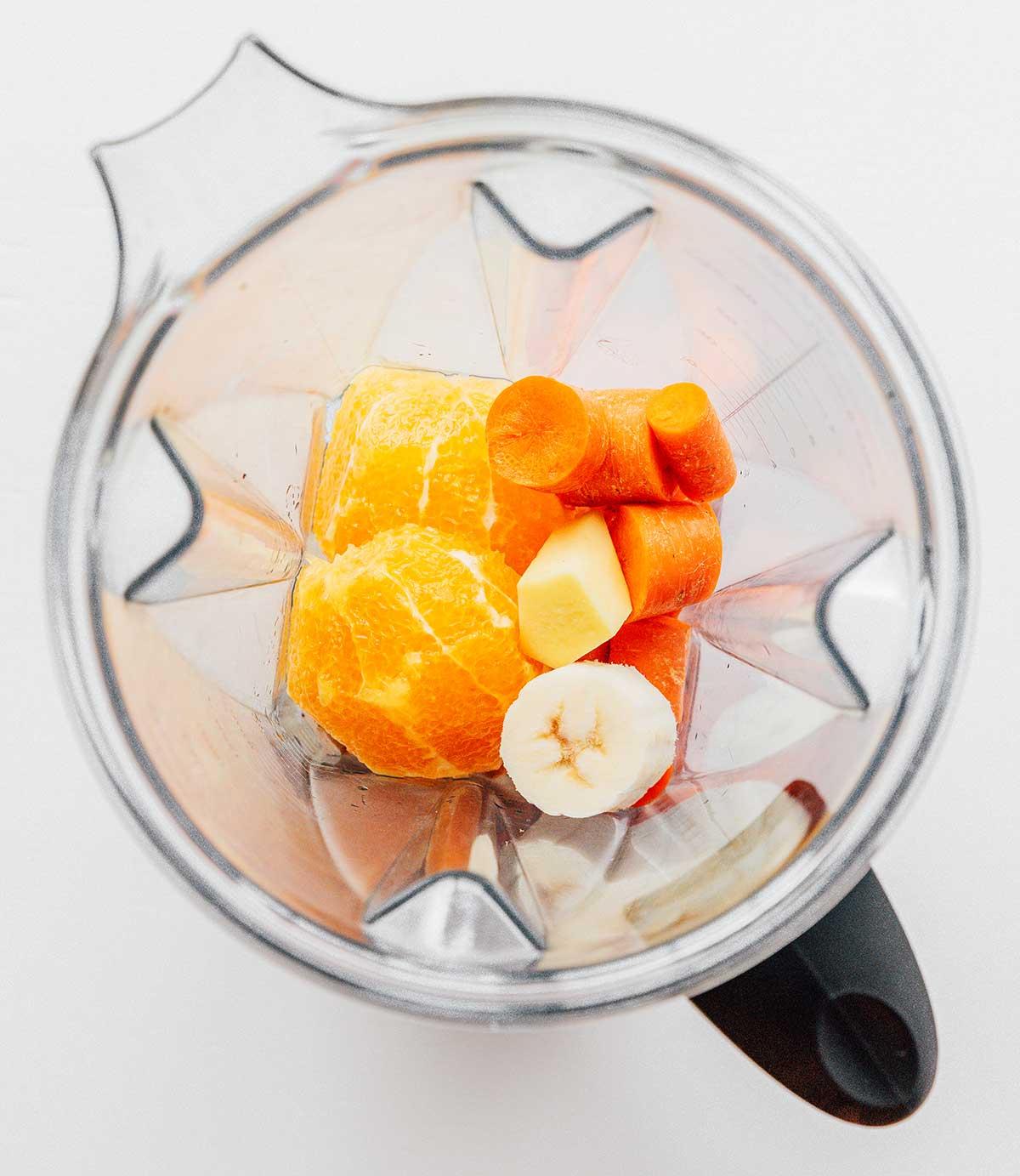 Ein Mixer gefüllt mit geschnittenen Bananen, geschnittenen Karotten, gehackter Ananas und 2 Hälften einer Orange