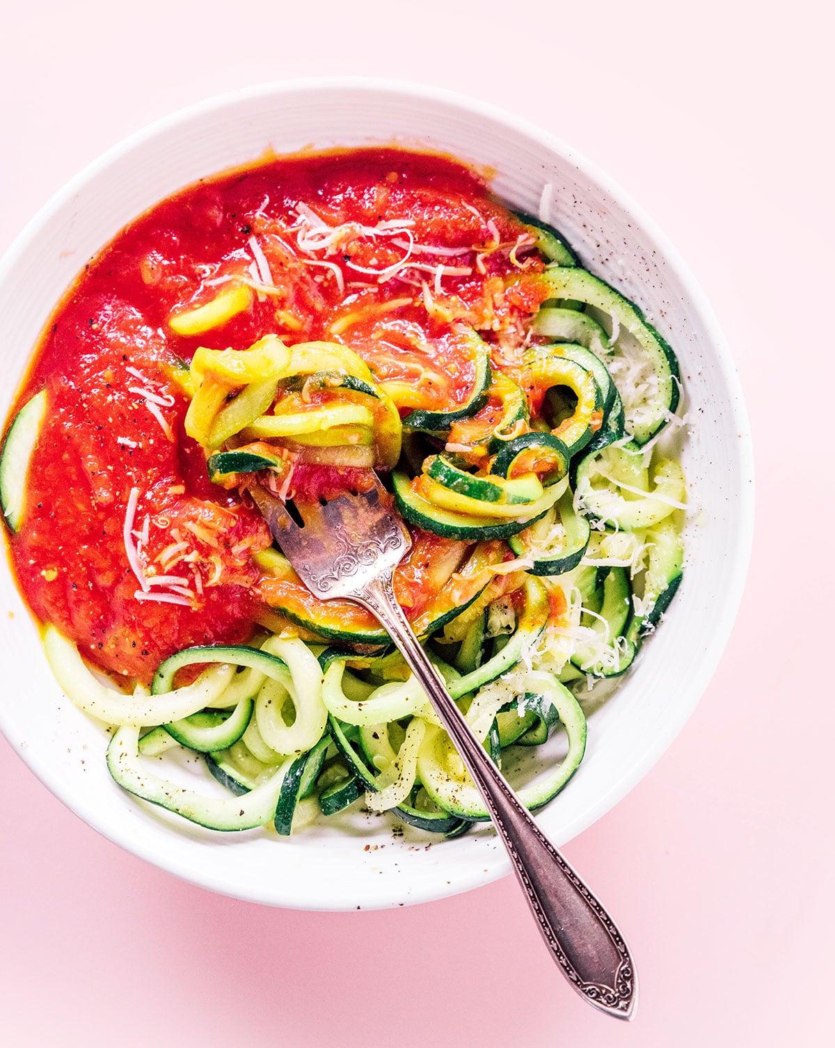 Zucchininudeln zubereiten 5 Spaghetti-Zucchini-Nudeln mit roter Soße, Parmesankäse und einer Gabel in einer weißen Schüssel.