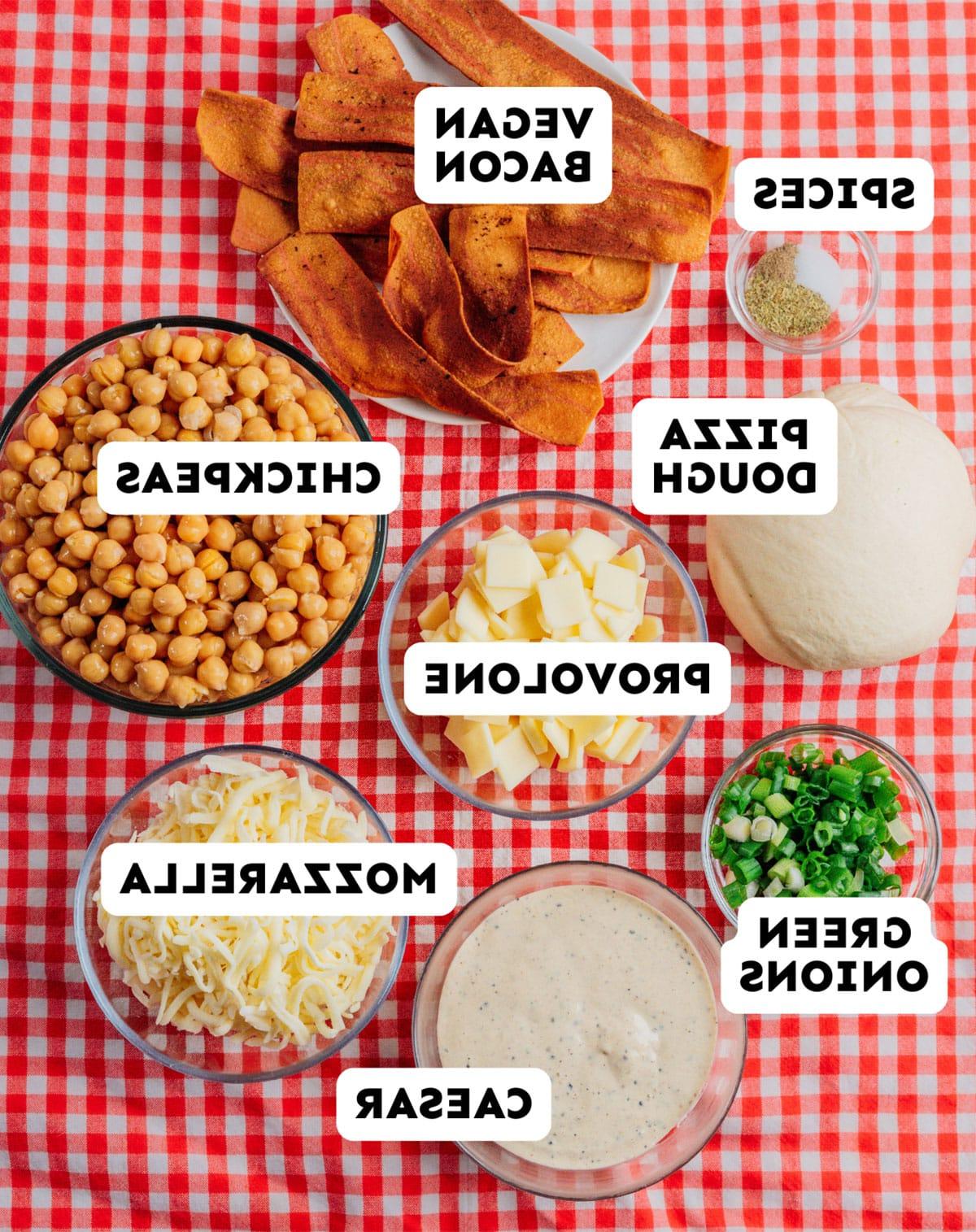 Vegetarischer Costco „Hähnchen“-Auflauf (Mit Kichererbsen!) 5 Verschiedene Pizzazutaten, beschriftet auf einer rot karierten Tischdecke: veganer Speck, Gewürze, Pizzateig, Provolone, Kichererbsen, Frühlingszwiebeln, Mozzarella und Caesar-Dressing – perfekt für die Kreation einer vegetarischen Costco Chicken Bake Variation.