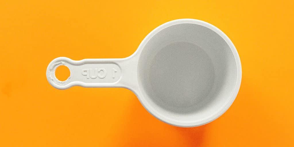 Wie viele Esslöffel sind in einer Tasse? (+ Umrechnungshilfe!) 5 Ein leerer weißer Plastik-Ein-Tasse-Messbecher auf einer orangefarbenen Oberfläche.