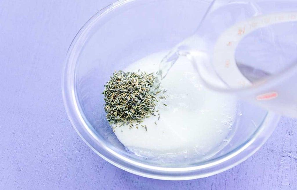 Einfache Lavendel-Zitronenlimonade (Saft oder Slush!) 5 Eine transparente Glasschüssel, gefüllt mit Lavendelblüten, Zucker und heißem Wasser, bereit zum Ziehenlassen, steht auf einer hellen, violetten Oberfläche.