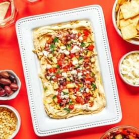 **10 Arten, Hummus zu essen (+ Rezepte!)** 5 Rechteckige weiße Platte mit einem geschichteten Hummus-Dip, garniert mit gehacktem Gemüse und Kräutern. Umliegende Schüsseln enthalten Pitachips, Oliven, Feta-Käse und Pinienkerne. Gläser mit Zitronenwasser stehen in der Nähe.