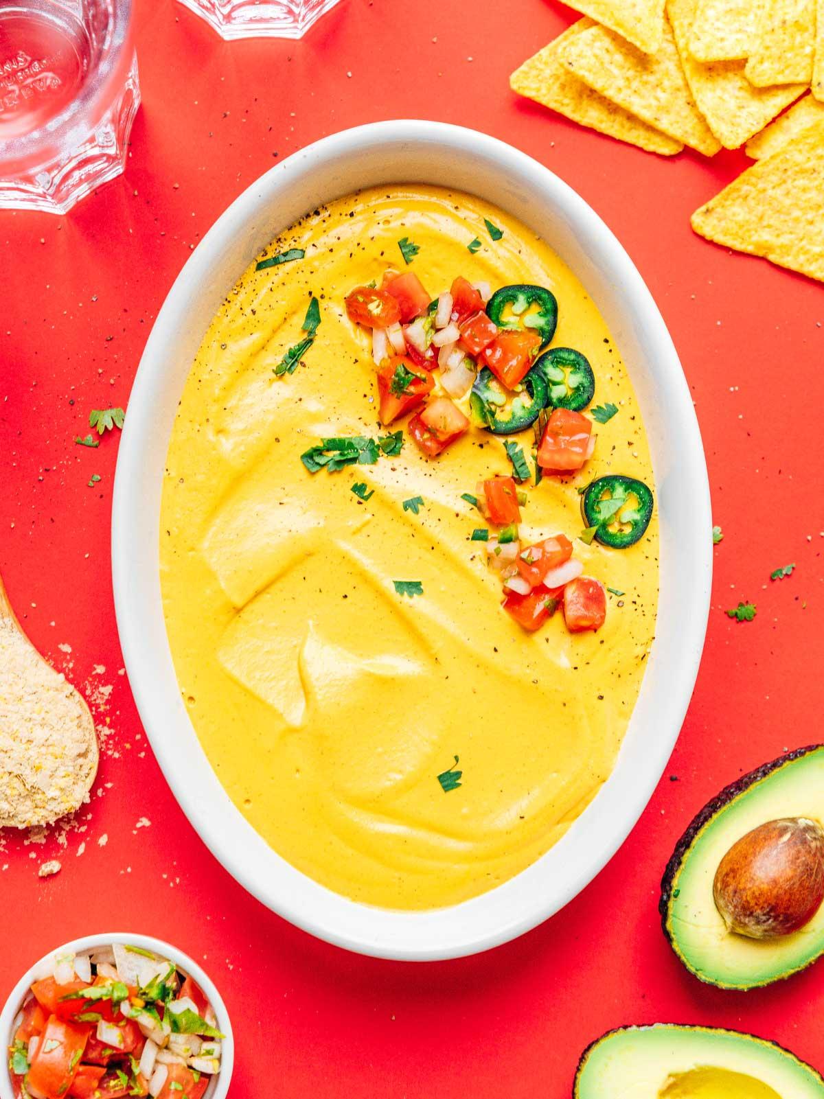**10-Minuten-Rezept für veganen Queso** 6 Eine weiße Schüssel gefüllt mit cremigem veganem Queso, garniert mit frischen Jalapeños, Tomatenwürfeln und Koriander, umgeben von mexikanischen Taco-Zutaten.