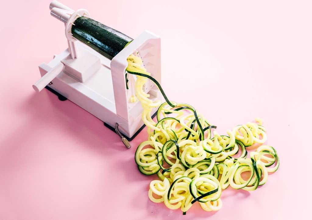 Zucchininudeln zubereiten 6 Ein Spiralschneider mit einer halben Zucchini darin und der anderen Hälfte, die durch den Spiralschneider geschoben und auf einem rosa Hintergrund zu Spaghetti-Zoodles verarbeitet wurde.