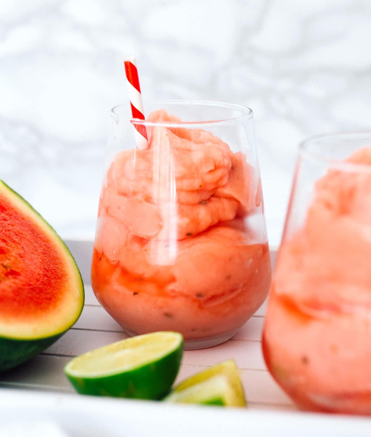 Gefrorener Wassermelonen-Daiquiri 6 Wassermelonen-Daiquiri in einem Glas mit rot-weißem Strohhalm und Wassermelonen- und Limettenscheiben daneben.