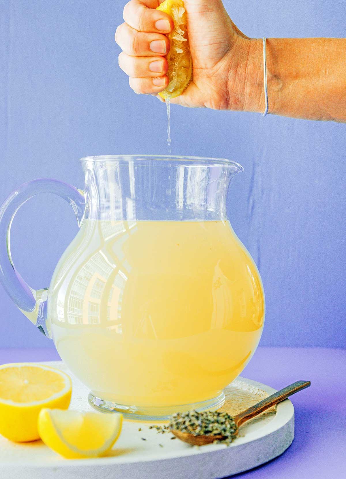 Einfache Lavendel-Zitronenlimonade (Saft oder Slush!) 6 Eine Hand presst frischen Zitronensaft in einen Krug mit selbstgemachter Lavendel-Zitronenlimonade, daneben stehen ganze und halbierte Zitronen.