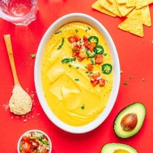 **10-Minuten-Rezept für veganen Queso** 7 Ovale Schüssel mit gelbem Käse-Dip, garniert mit gehackten Tomaten, Koriander und Jalapeños, umgeben von Tortillachips, Avocadohälften, einer kleinen Schale Salsa, Seitan-Flügeln und einem Holzlöffel mit Gewürzen.