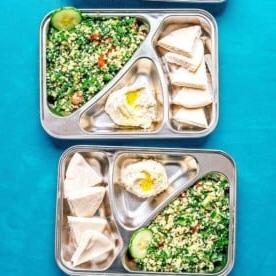 **10 Arten, Hummus zu essen (+ Rezepte!)** 7 Drei Metalltabletts mit unterteilten Fächern präsentieren einen lebendigen Tabouleh-Salat, dazu cremigen, mit Olivenöl beträufelten Hummus, Pitabrot und erfrischende Gurkenscheiben, alles vor einem atemberaubenden türkisfarbenen Hintergrund.