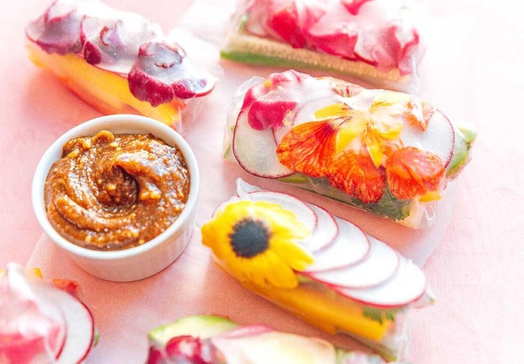 Essblumen-Frühlingsrollen 8 Essbare Blüten-Sommerrollen auf rosafarbenem Hintergrund neben einer Schale mit Mandelbutter-Dip-Sauce