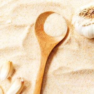 Wie man Knoblauchsalz herstellt 8 Ein Holzlöffel liegt auf einem Haufen Knoblauchpulver, daneben eine ganze Knoblauchknolle und drei Knoblauchzehen, die auf die perfekte Geschmacksnote hinweisen, die nur eine Prise Knoblauchsalz bieten kann.
