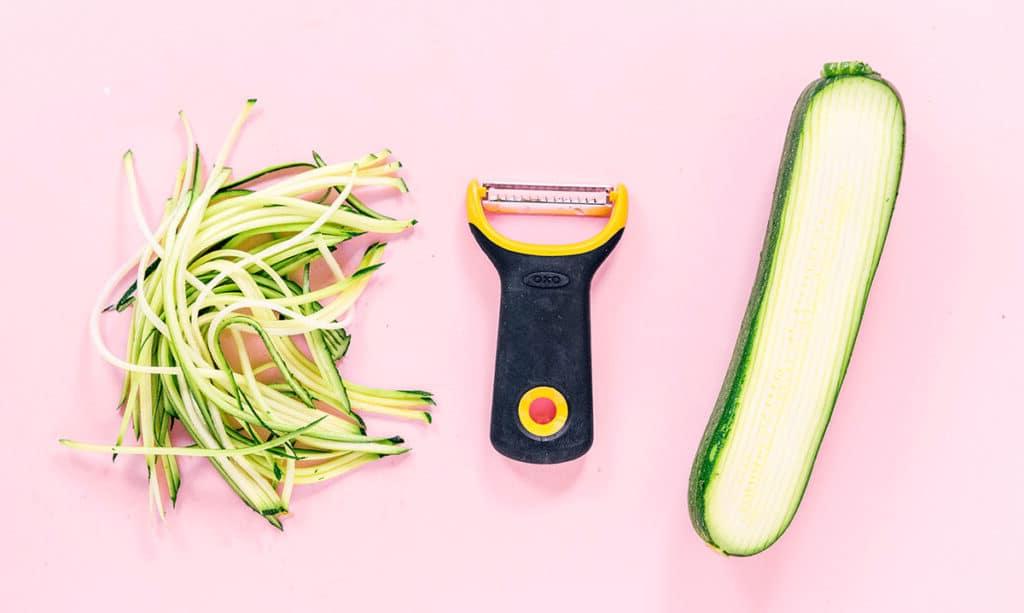Zucchininudeln zubereiten 8 Eine halb geschnittene Zucchini, ein schwarzer und gelber Julienne-Schneider und ein Stapel engelshaariger Zucchini-Nudeln, die auf einem rosa Hintergrund angeordnet sind.