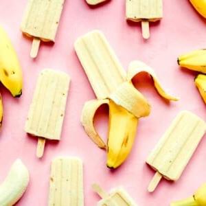 Bananen-Eis am Stiel aus 3 Zutaten 8 Bananen-Popsicles und ganze Bananen bilden ein spielerisches Muster auf einem leuchtend pinken Hintergrund.