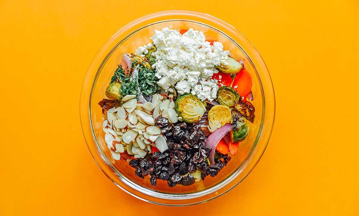 Geröstete Gemüse-Getreide-Bowls 8 Eine durchsichtige Glasschale, gefüllt mit Zutaten für eine geröstete Gemüse-Getreide-Bowl wie geröstetem Gemüse, Feta-Käse, Mandelblättchen und getrockneten Kirschen.